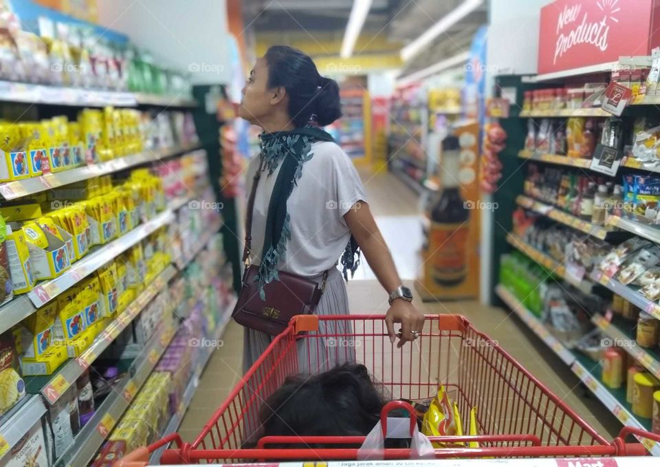 mother was looking for items at the grocery counters while her curly daughter was sitting on the shopping cart.