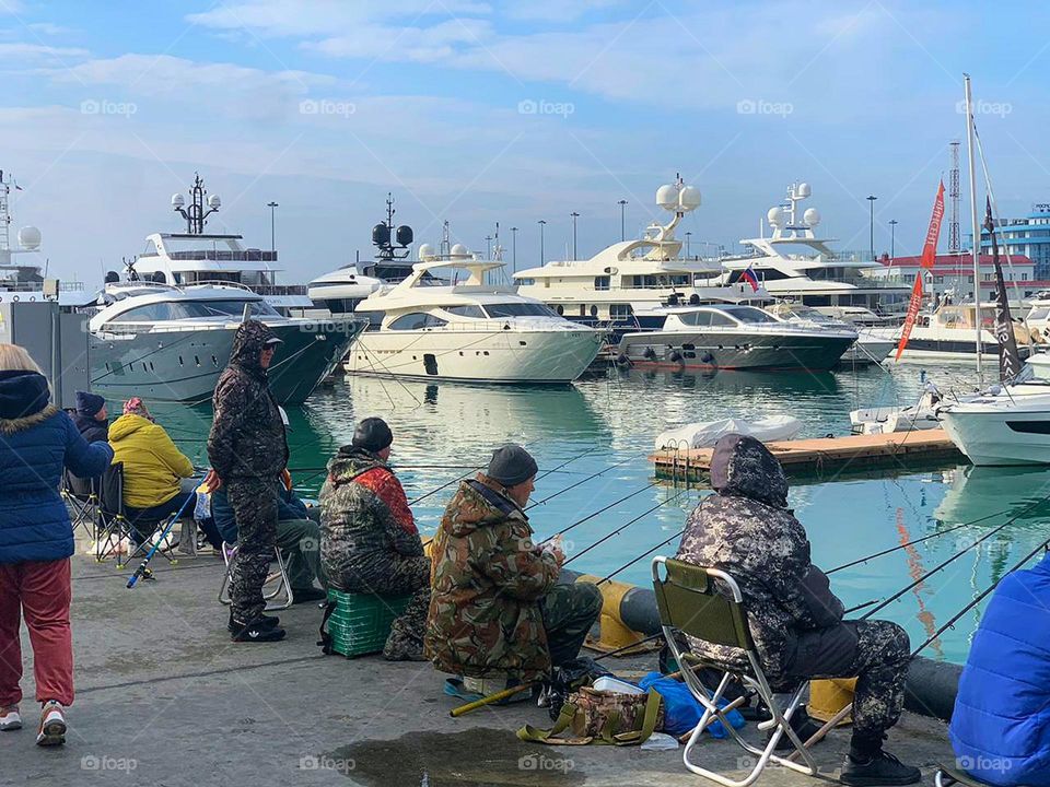 Fishermen sit on the pier.  White yachts stand in the blue sea