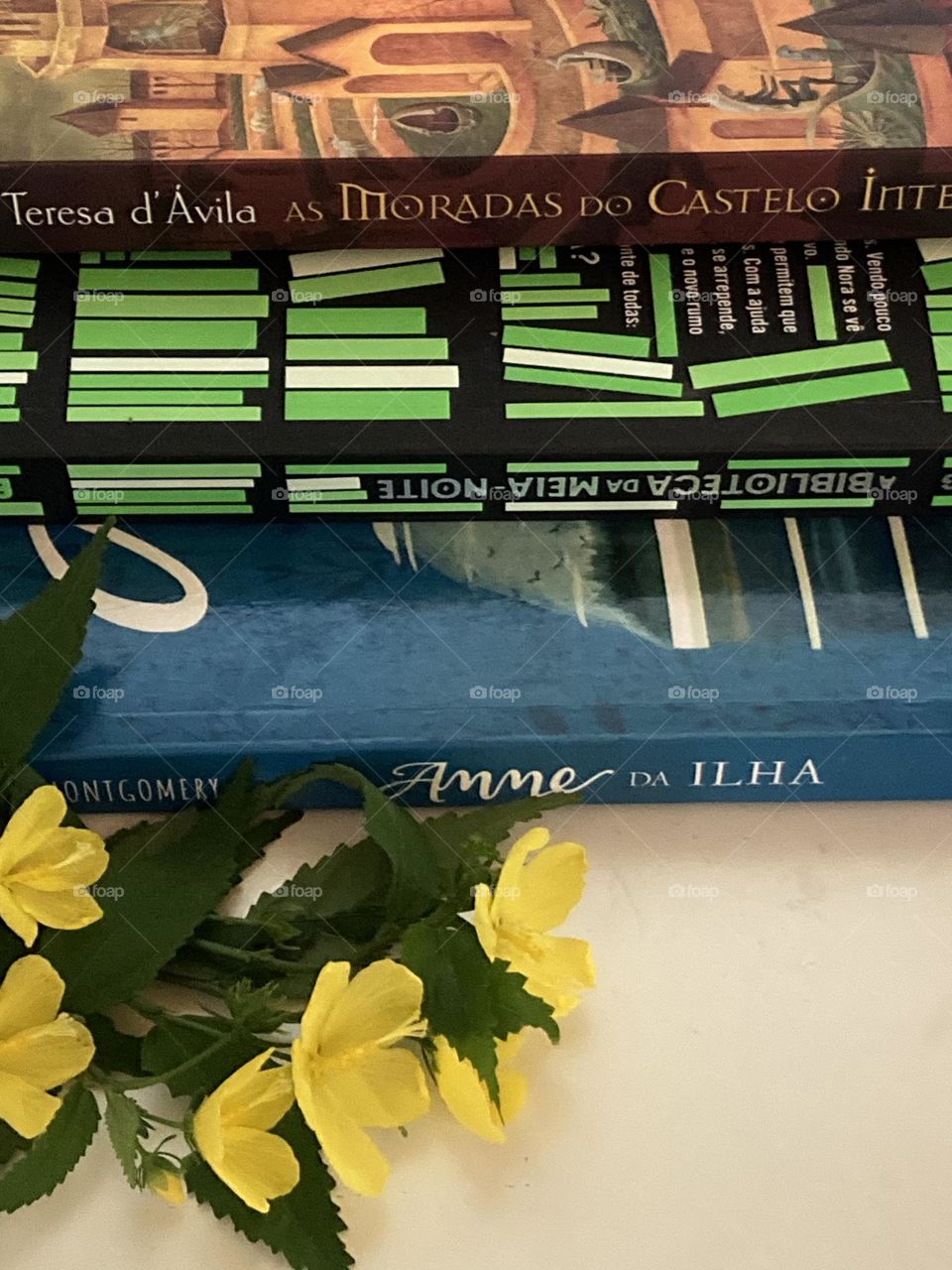 Mesa branca com alguns livros de leitura e ao lado buquê de flores amarelas enfeitando a cena.