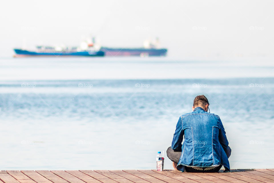 Man Wearing Denim Jeans Clothes Sitting On A Dock And Looking At Blue Sea
