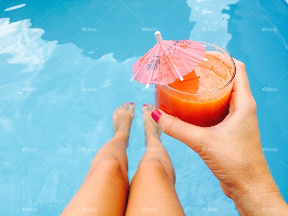 Woman hand with pink nail polish holding glass of orange juice with umbrella over the pool blue water