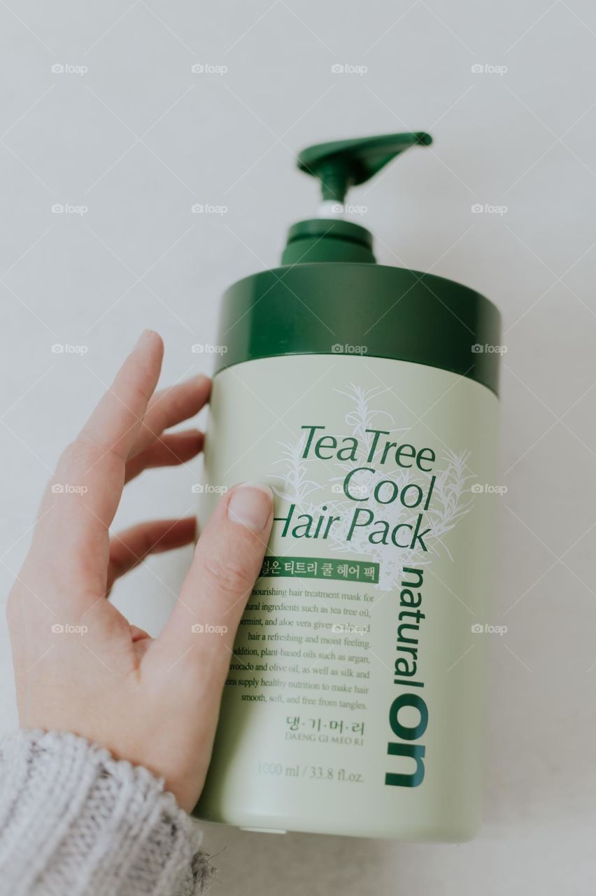 The hand of a young Caucasian woman touches a large green bottle with a dispenser of a professional hair mask brand natural ON lying on a light background, flat lay close-up.