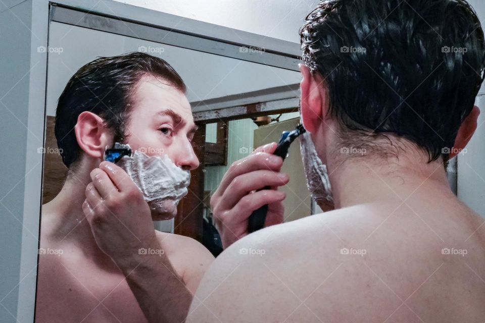 A man shaves while looking in the bathroom mirror
