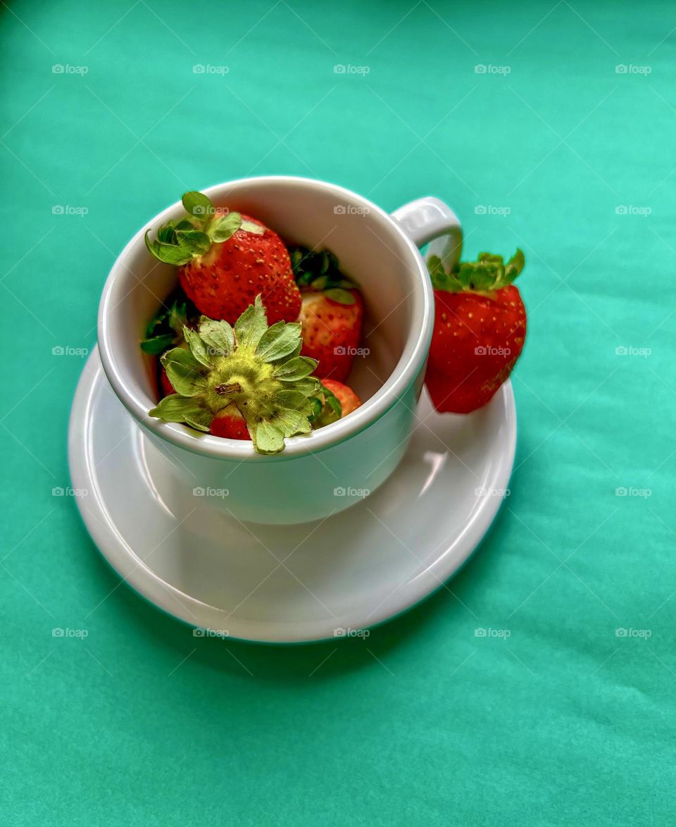 A white cup full of fresh and juicy strawberries, with a slice of strawberry on the side. The vibrant green background highlights the red color of the strawberries