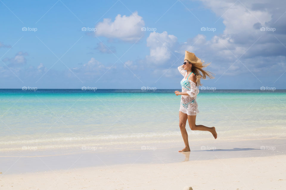 Happy young woman with long blonde hair in straw hat, sunglasses and white tunica running on the beach 