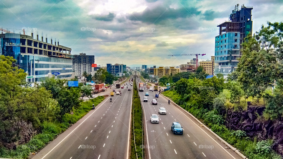 Indian road scenery captured from bridge. This photograph shows Indian road with traffic and it's roadside surrounding with trees, buildings and cloudy sky.