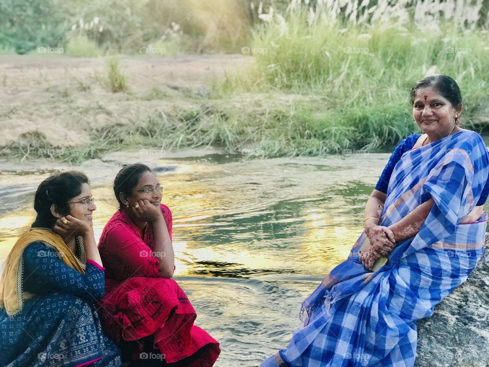 Two girls sitting on rock in the middle of river and looking towards a smiling woman and a beautiful sunset at back 