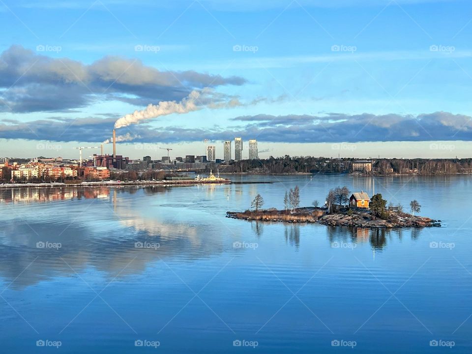 Tranquil scene with Helsinki coastline cityscape with smoking pipe, little island with trees and small wooden cottage and clouds reflecting in water 