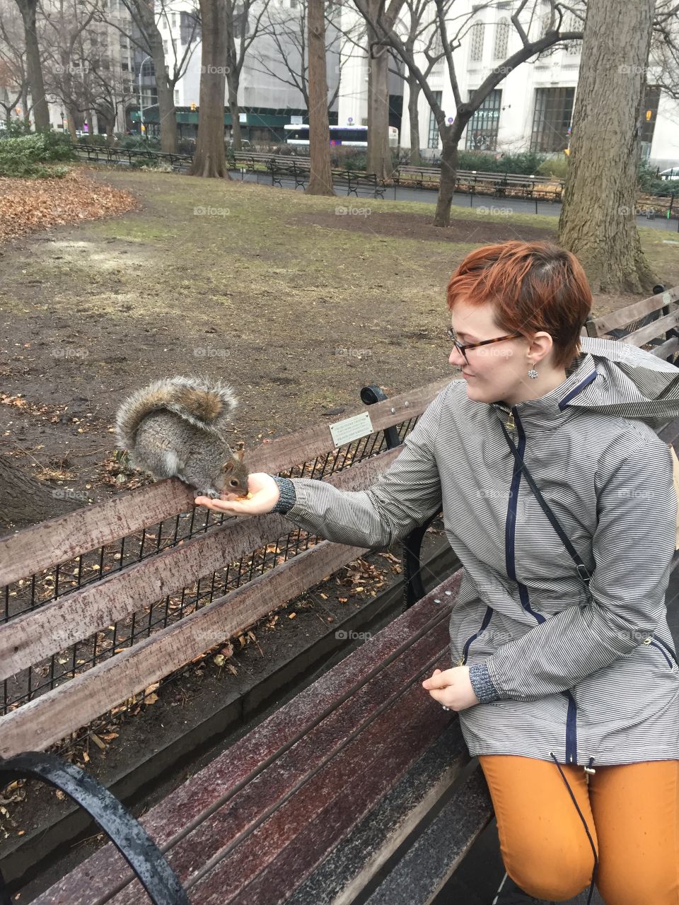 Young woman with short red hair and glasses wearing jacket and feeding squirrel in the park