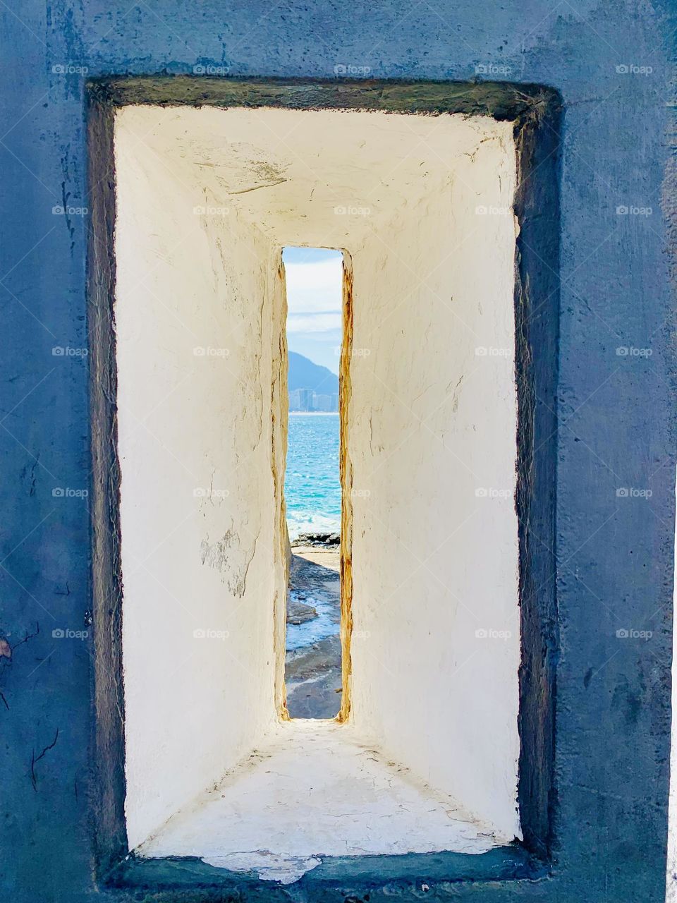 A picturesque view of the marine landscape captured through a rectangular concrete frame, displaying a distant horizon with calm blue waters and a mountainous setting, evoking a feeling of peace and serenity.