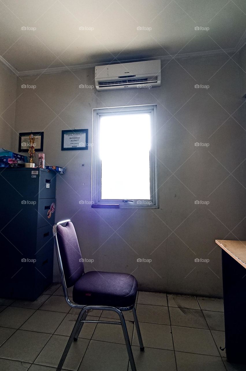 corner of the room with a chair, table, filing cabinet, and a window that emits bright light