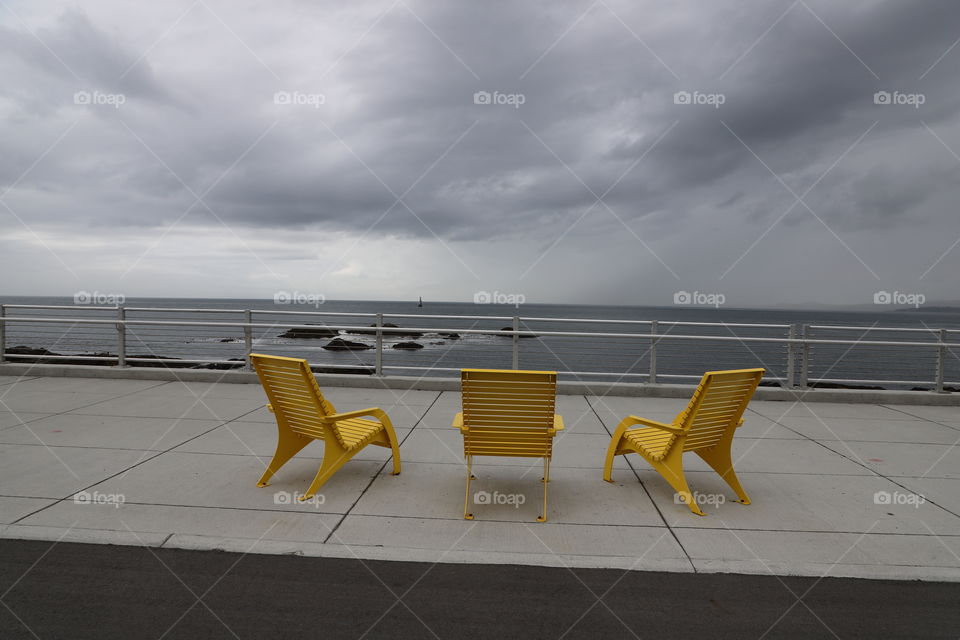 Yellow lounge chairs on a sidewalk facing the grey ocean and cloudy sky 