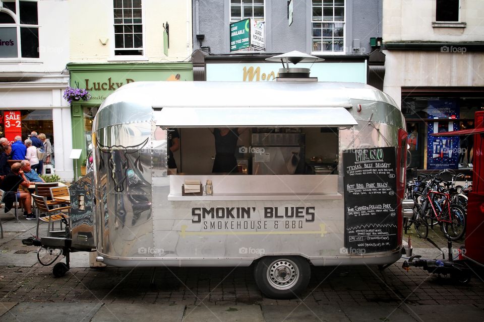 A shiny, silver BBQ van in a street market selling grilled meat and street food.