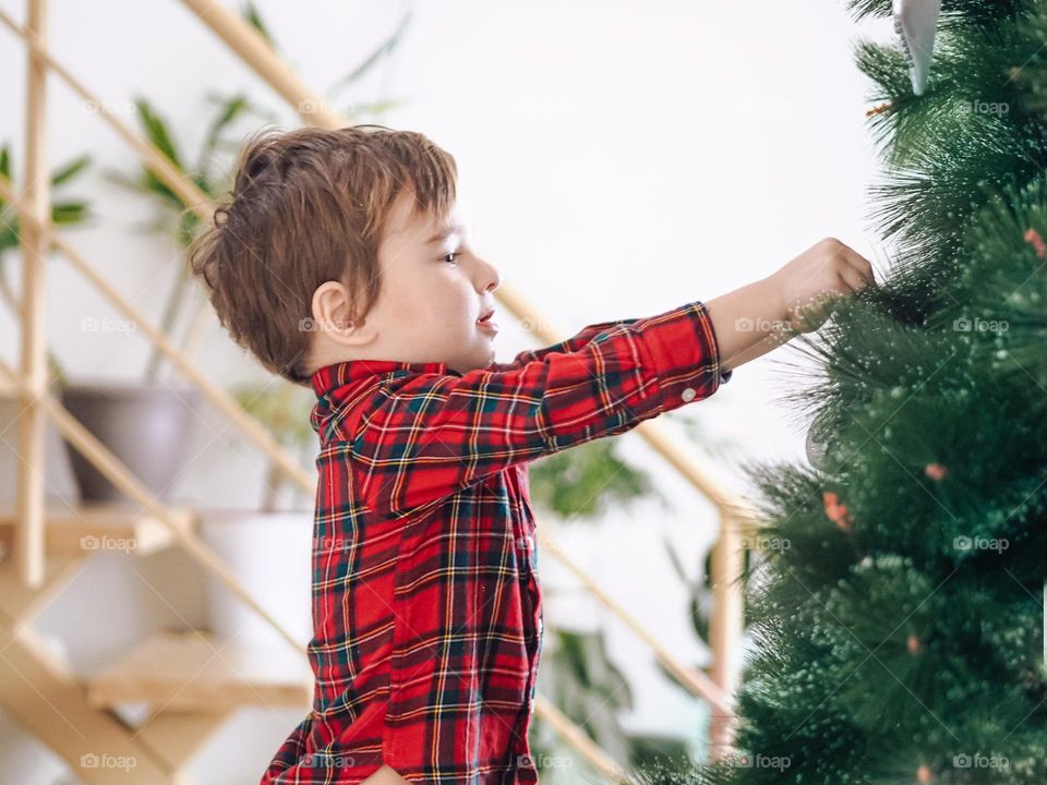 Cute toddler boy in plaid shirt decorating Christmas tree 
