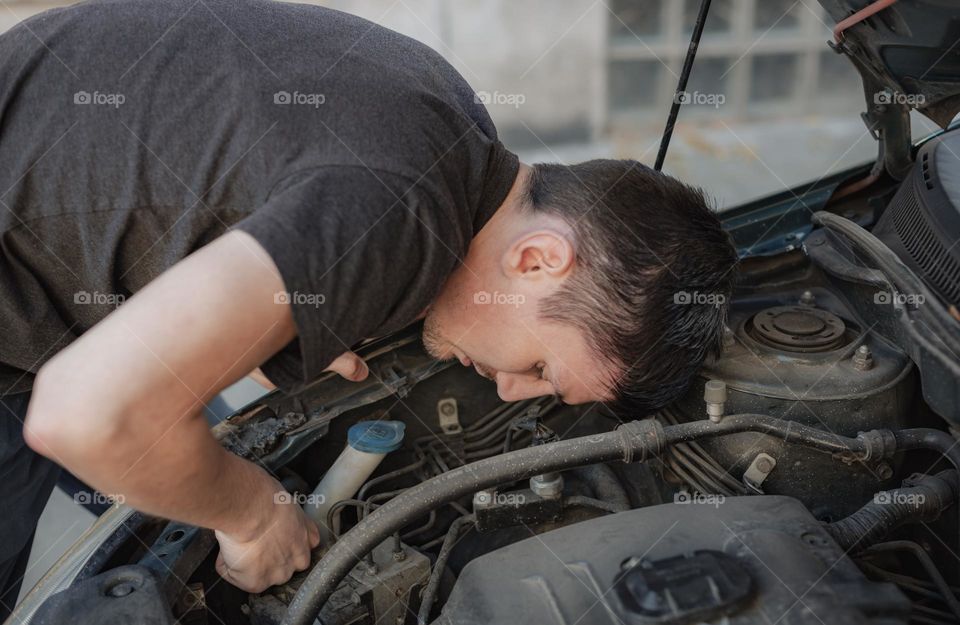 One young caucasian man is pulling out a burnt out light bulb from the left front headlight while standing bent over during the day on the street, side view close up. Car repair concept.
