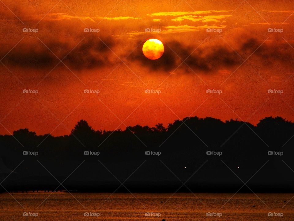 A reddish orange sunrise over the lake at Cranes Roost Park. The sun looks like it is sitting on some dark clouds.