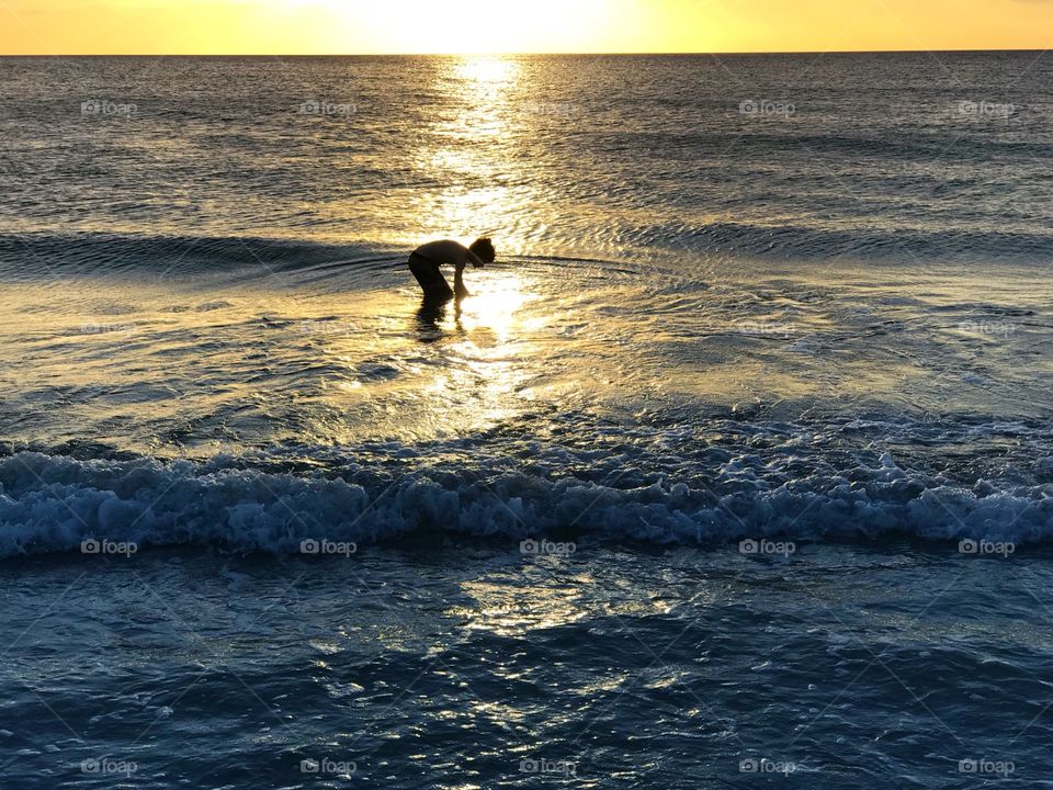 Small boy joyfully playing in the ocean in the reflection of a bright yellow sunset.