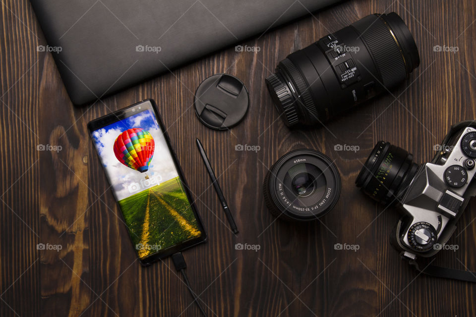 Flatlays of the notebook,  photo lens, camera , smartphone.  display of the mobile phone shows Foap picture.  my favorite gadgets makes me money and gives pleasure.  it is an opportunity to become a part of the Art