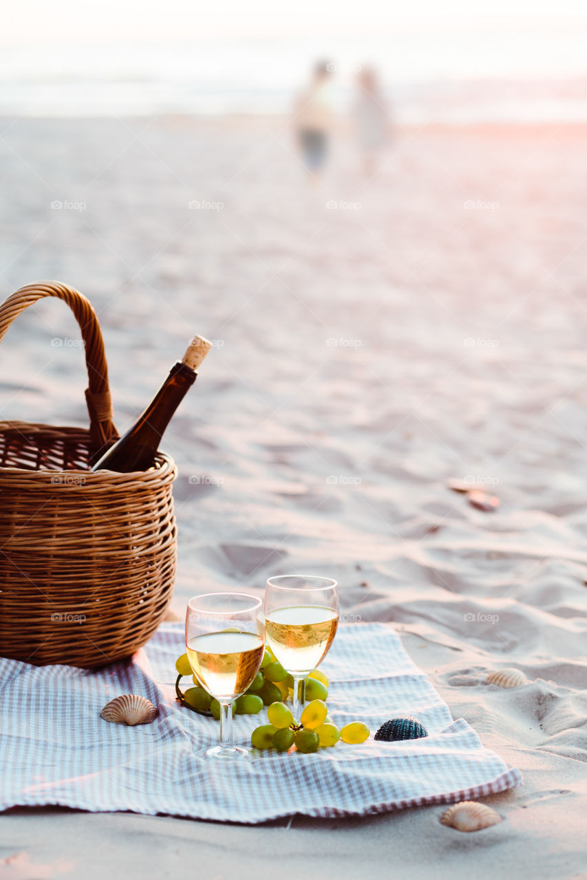 Two wine glasses with white wine standing on sand, on beach, beside grapes and wicker basket with bottle of wine. Young couple standing in the sea in the background