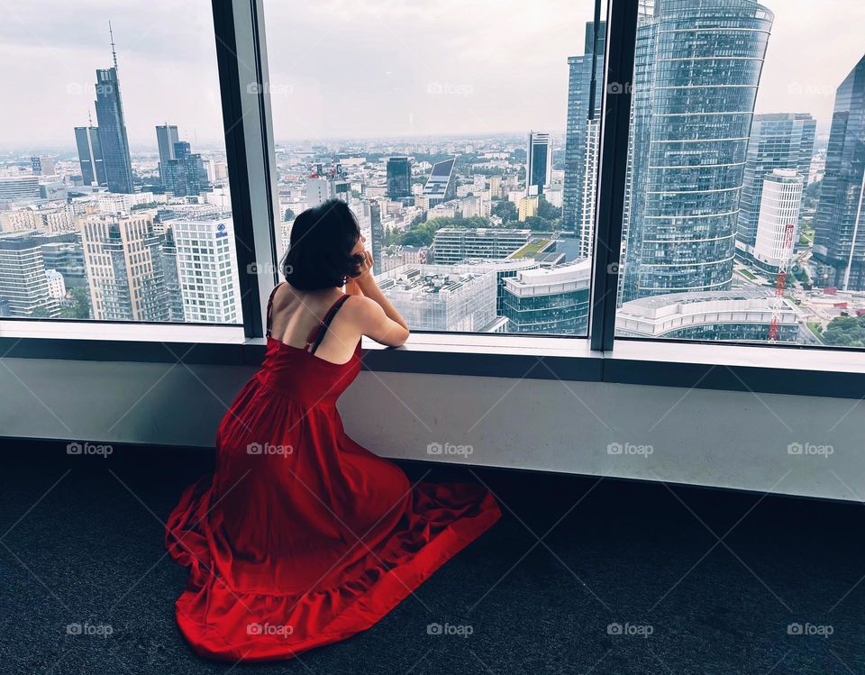 Woman in red dress in front of window with view on skyscrapers 