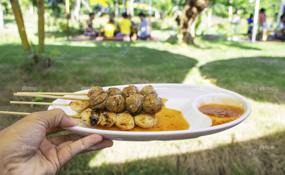 Grilled pork and beef meatballs with seafood sauce on white plastic plate in hand Background blurry Tourists on the lawn.