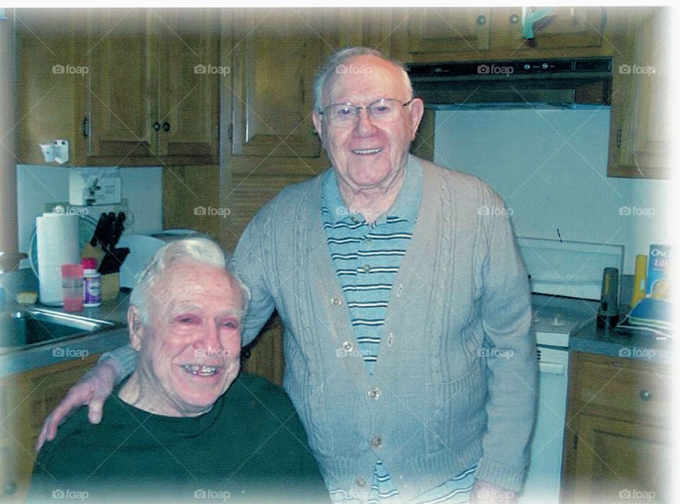 Two elderly men, also good old friends posing for a photo while visiting each other in a kitchen.
One older man standing, the other sitting. Both are senior citizens, grandfathers, grandparents & relatives.