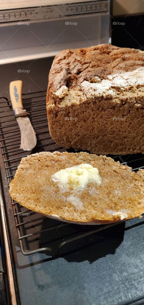 Hot buttered slice of homemade Cracked Wheat bread 1.5 pound loaf. Cut slice of hot bread with butter melting into it. Bread loaf cooling on rack with butter knife to the side.