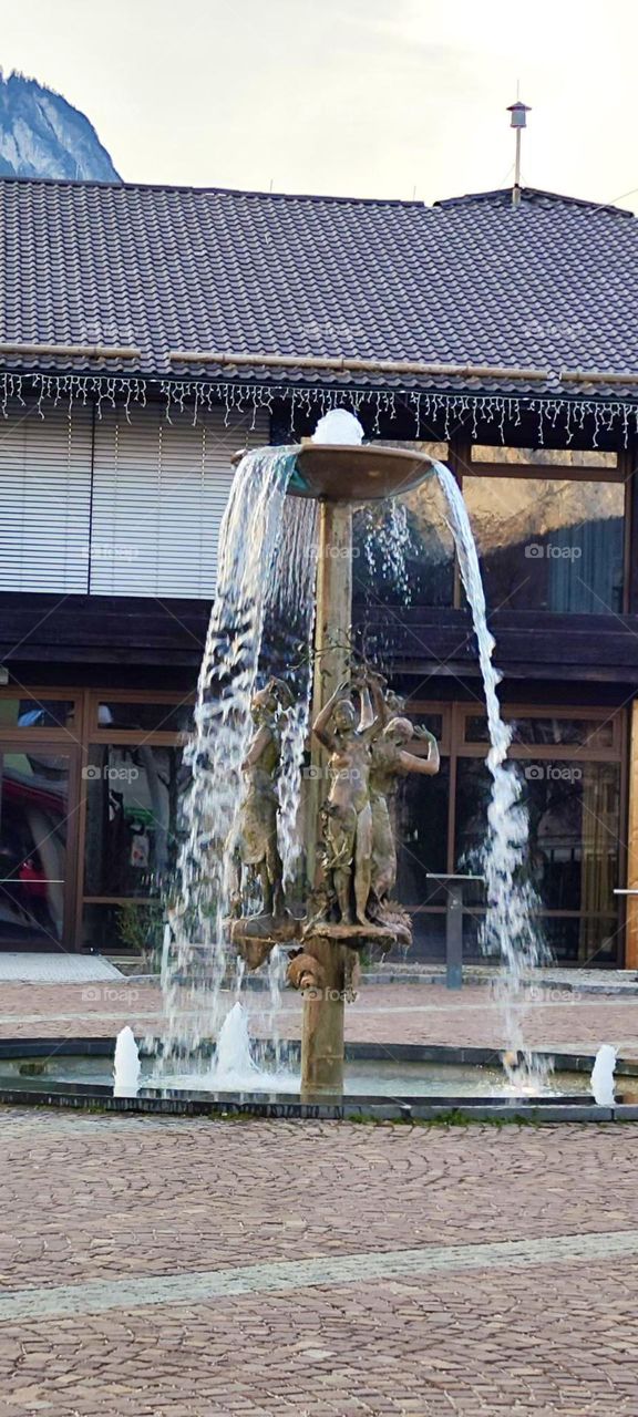 A tall artful fountain w sculptures of women raising their hands & arms seemingly reaching for the refreshing clean mountain water can be found in the picturesque little town of “Garmisch Partenkirchen” in the Bavarian Alps. 2023. Hypnotic Productons