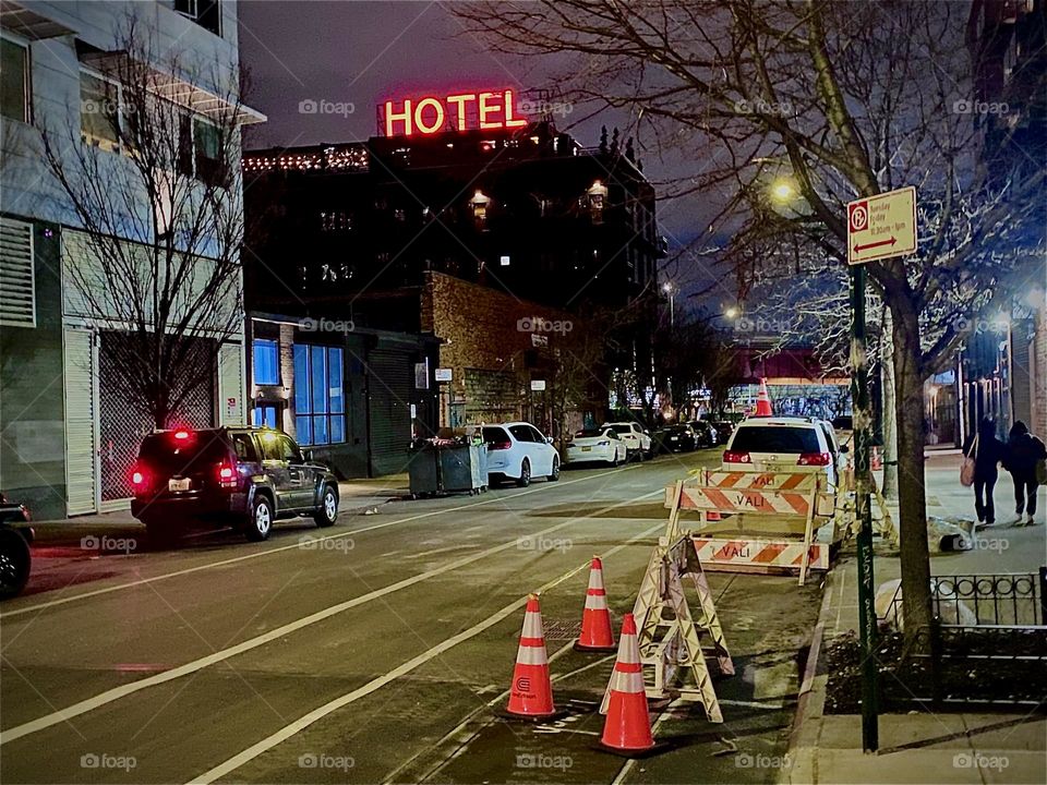 A view from a bit further back at the „BOXHOUSE Hotel“ walking down „Boxstree“ with a lit up bright red „HOTEL“ sign on top of the building. The street is illuminated brightly and a construction site is boarded off. 2023. Hypnotic Productions