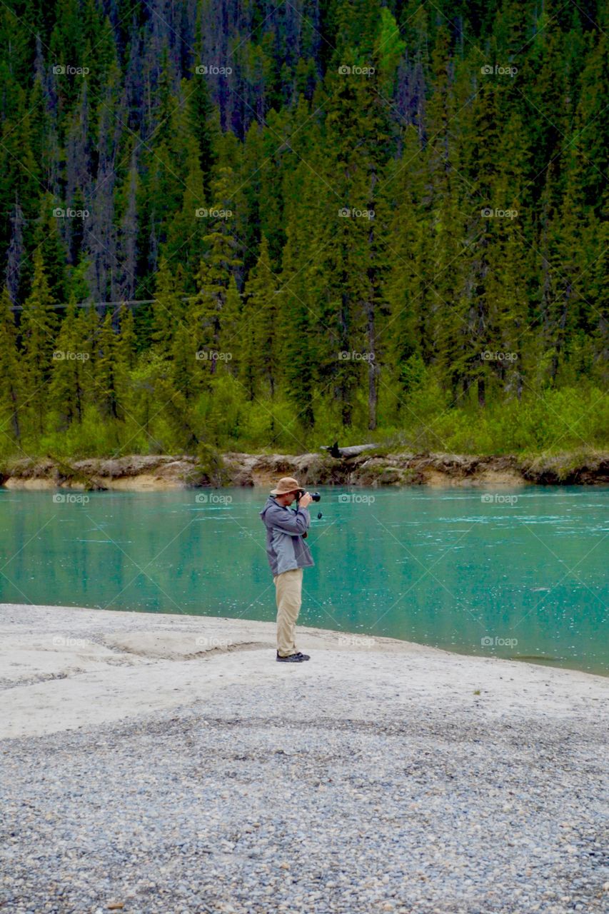 Male photographer by turquoise glacier fed river taking photographs with DSLR camera 