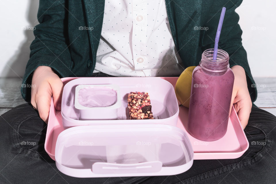 A schoolboy sits on the floor in a lotus position with a tray in his hands with a healthy snack, yogurt, a cereal bar, an apple and an oatmeal cocktail. Healthy school meals. College lifestyle