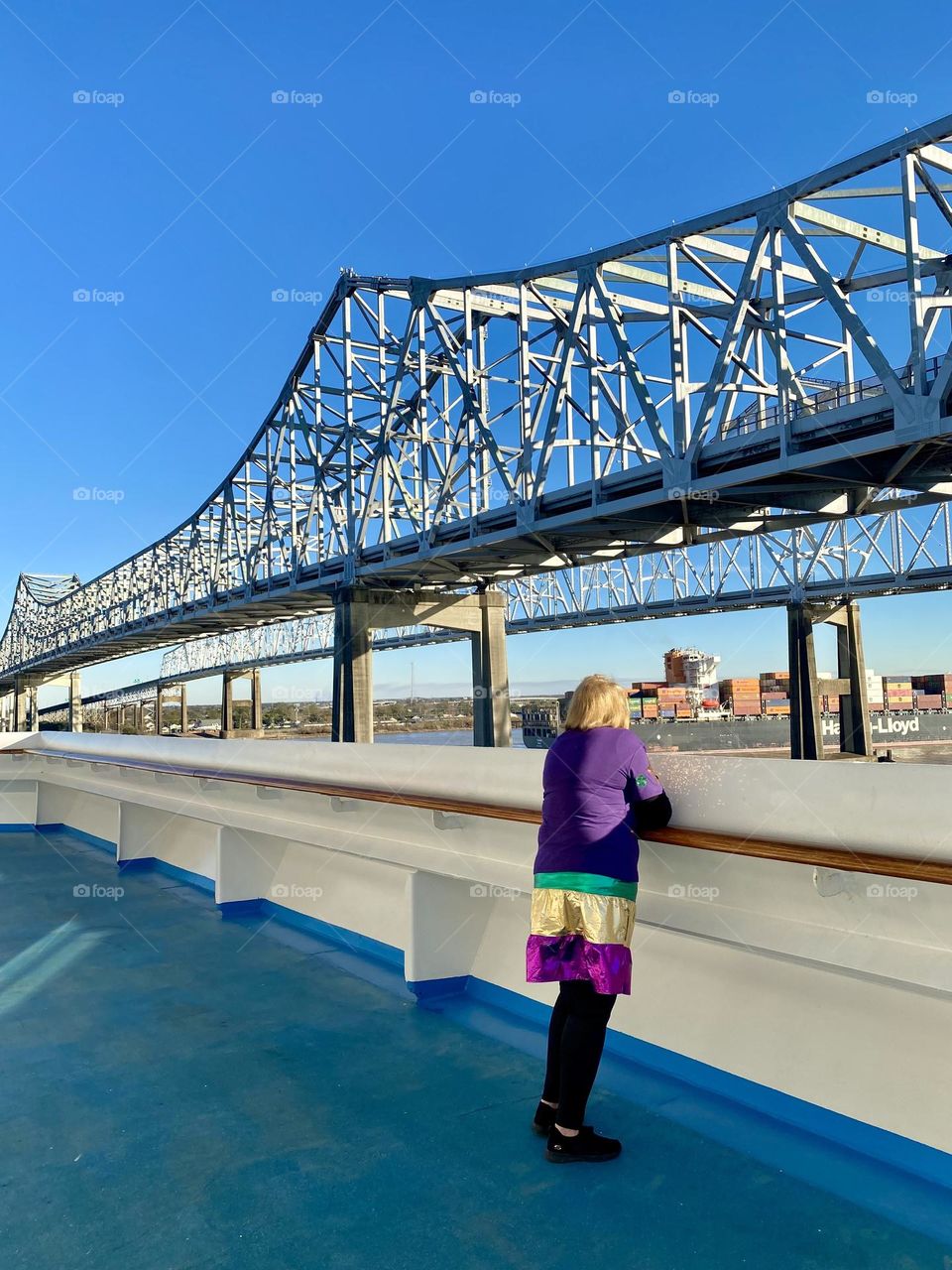 A lady wearing Mardi Gras colors standing at the railing on a cruise ship looking at the view of a bridge