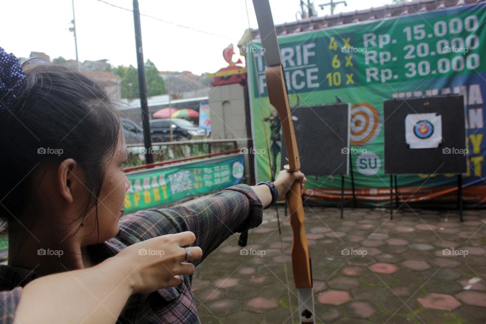 A young Asian woman in plaid shirt holding an archery and aiming at the arrow target in front of her, Semarang, Indonesia.