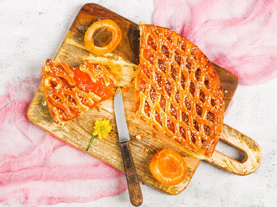 Beautiful appetizing cut piece of apricot pie with fresh apricots on a wooden cutting board with a pink gauze napkin lie on a light cement background, flat lay close-up.