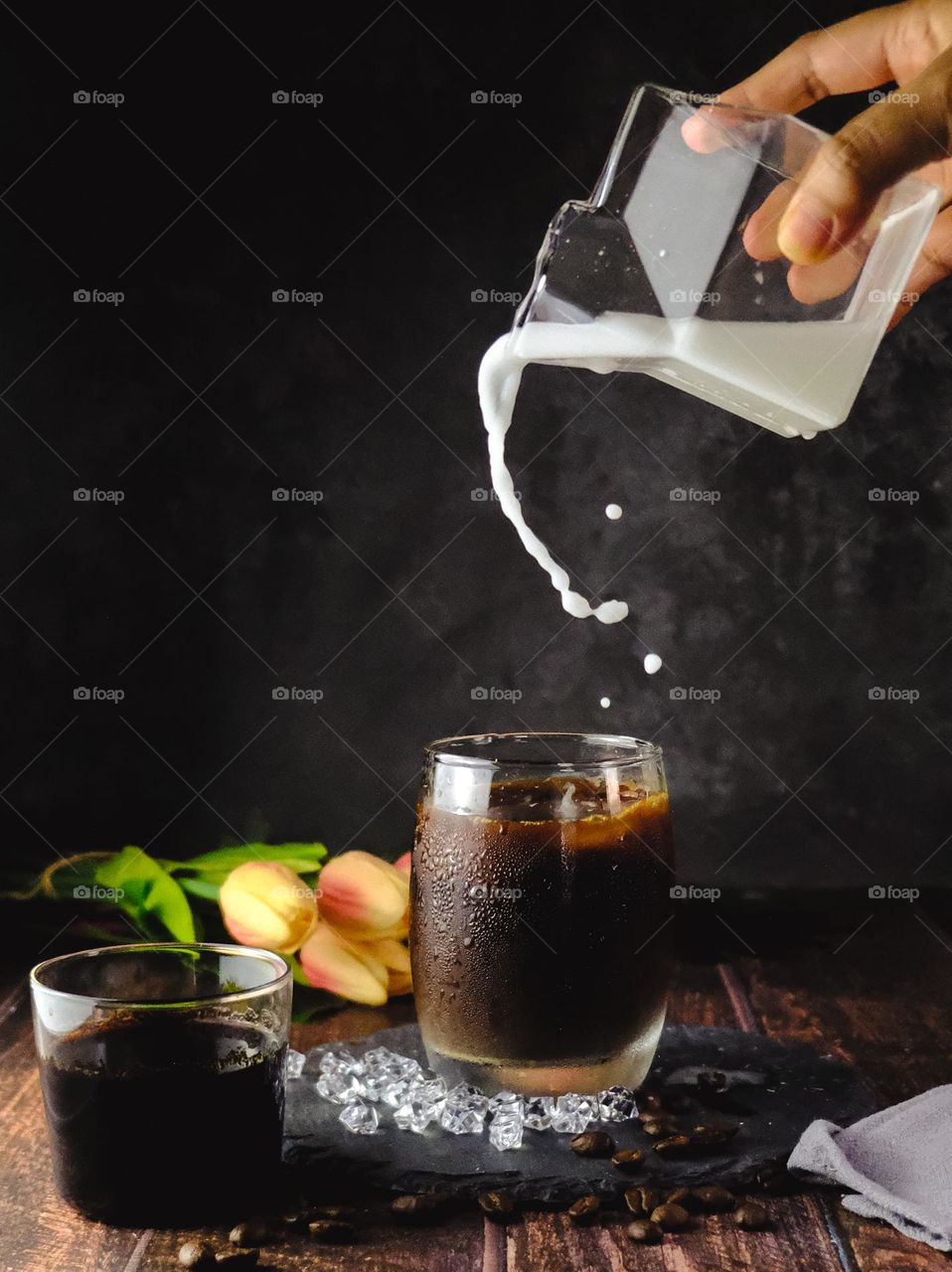 Woman pouring milk from glass jug into one of two glasses with espresso coffee on wooden stand with dark background.