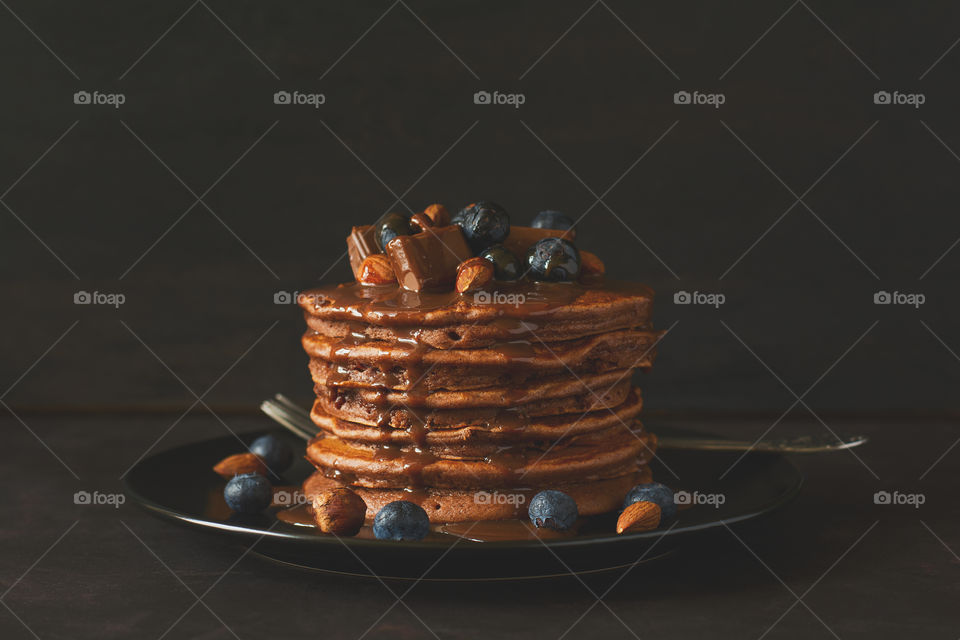 Stack of pancakes with chocolate sauce, blueberries and nuts on dark wooden desk