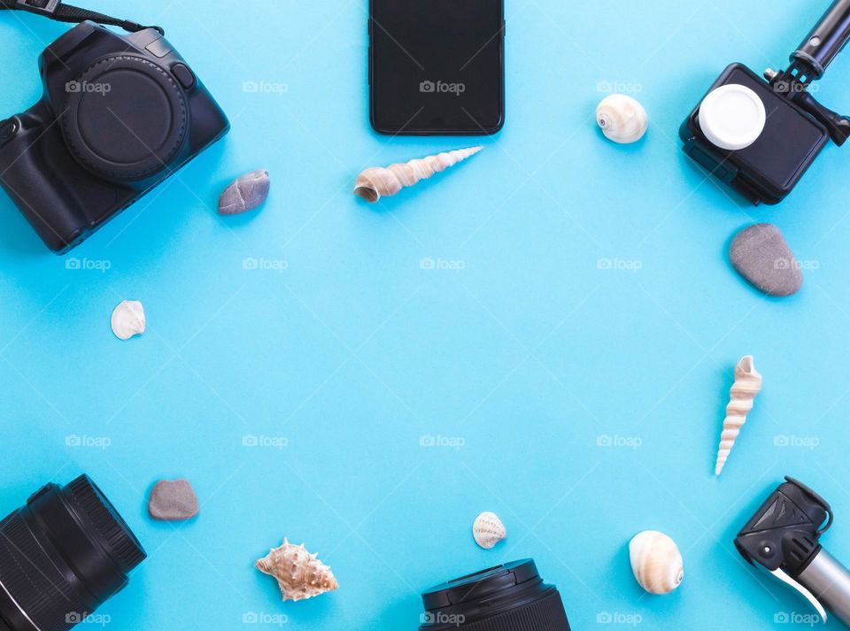 Photographer's set of camera, action camera,selfie stick, lenses,smartphone with black screen and shells with stones lie oval frame on blue background with copy space in the center,flat lay close-up.background with copy-space space,flat lay close-up.