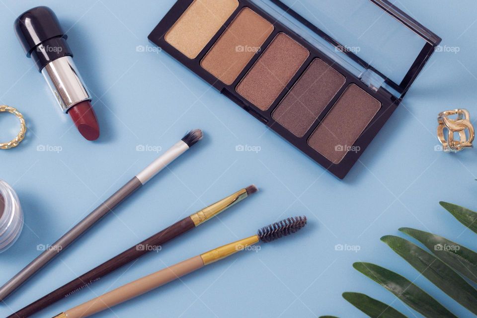 A pallet of nude eye shadows, makeup brushes, eye pencil, gold ring and a palm branch on a blue background, flat lay. Concept female cosmetics, beauty salon.