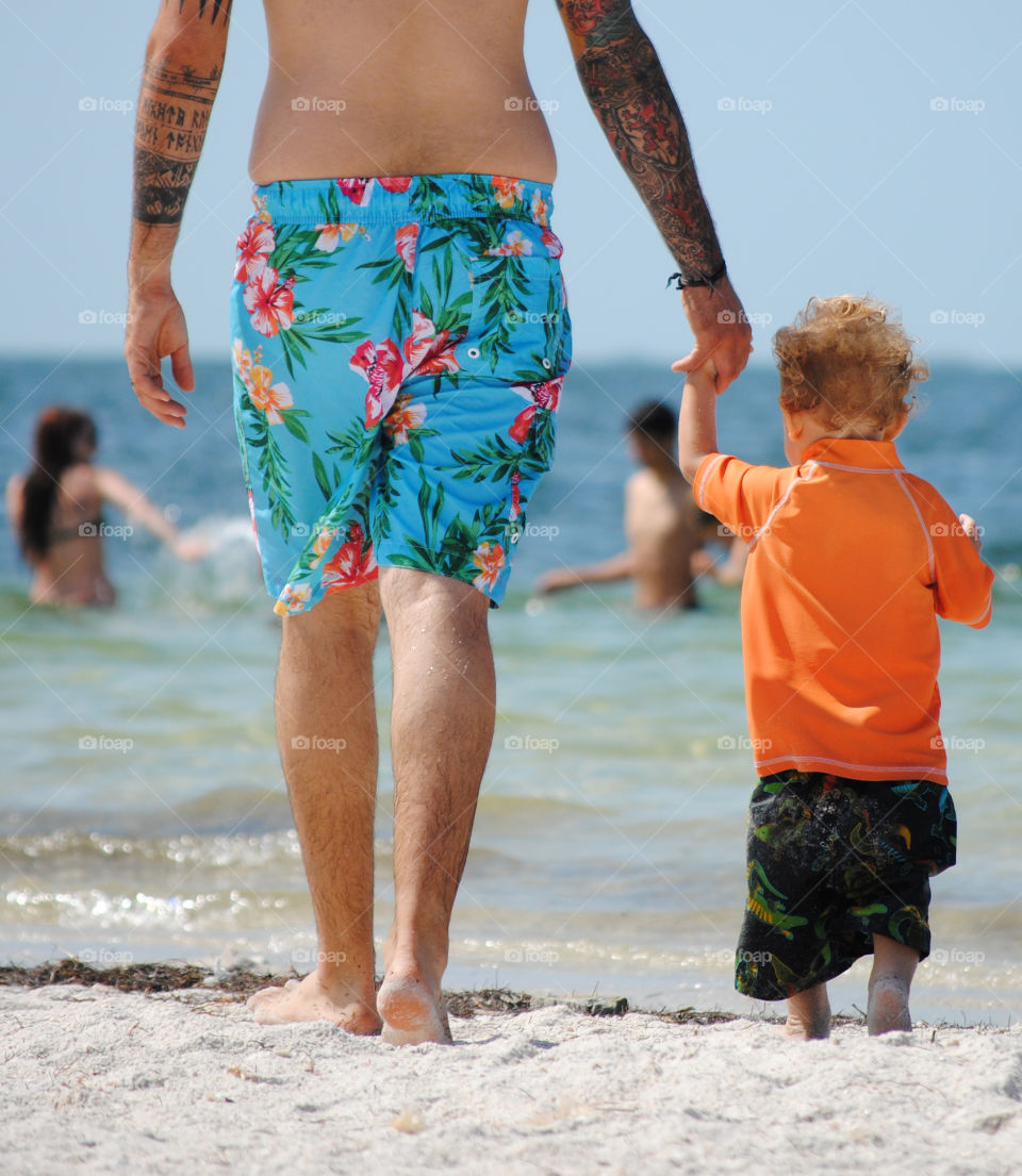 summer time fun for a toddler and his family at the beach as dad holds his hand to walk him across the sand to the water