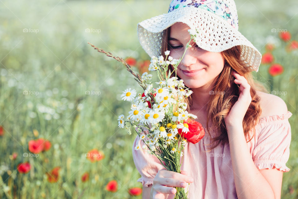 Beautieful young girl in the field of wild flowers. Teenage girl picking the spring flowers in the meadow, holding bouquet of flowers. She wearing hat and summer clothes. Spending time close to nature