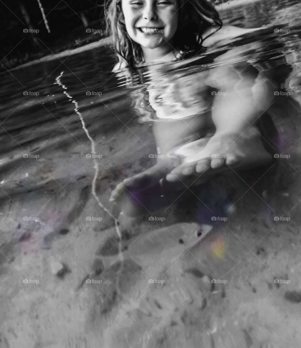 A girl sits in very clear water while a fish swims by her feet. Reflection