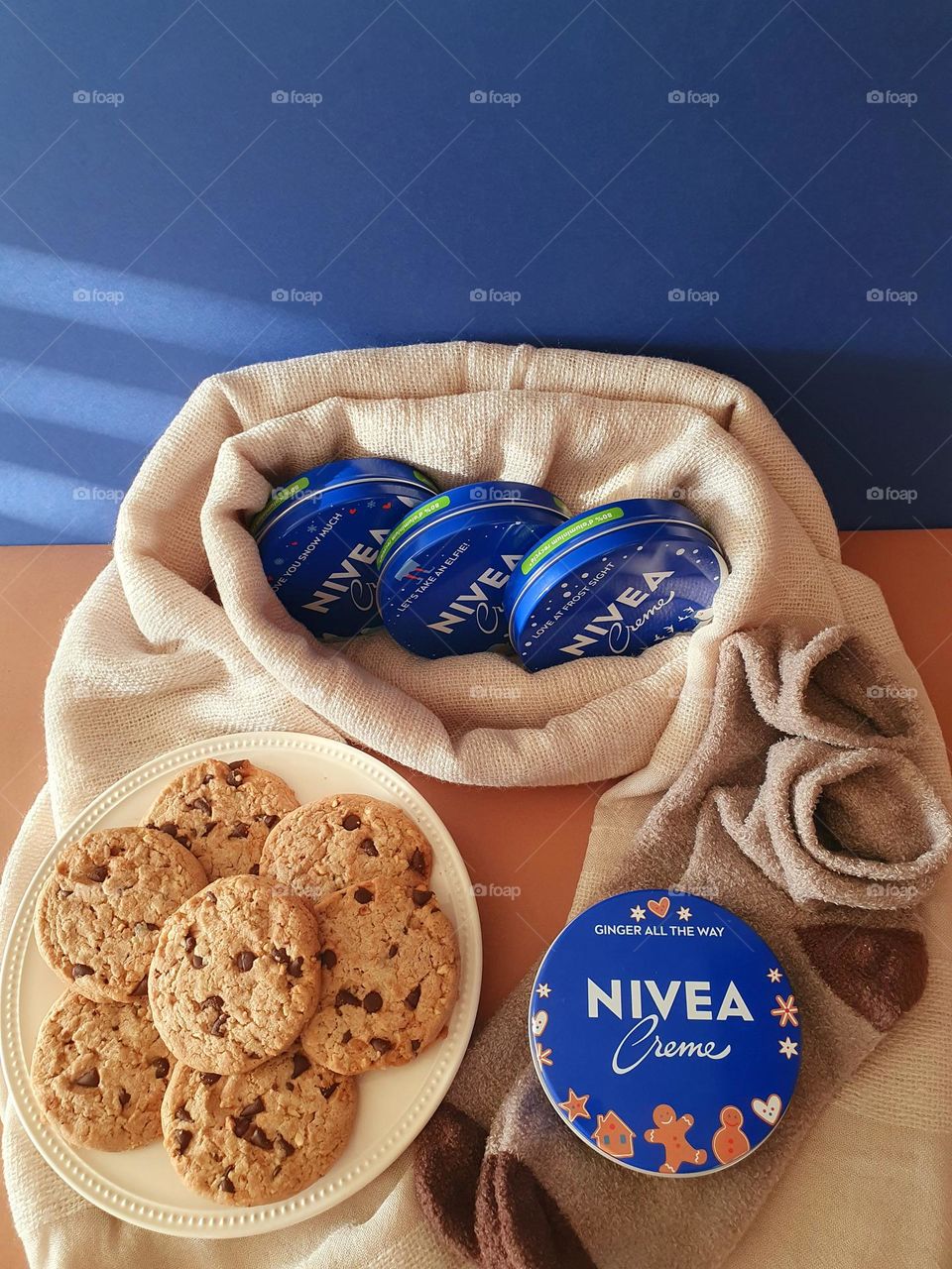 The 4 tins of Nivea Crème winter limited edition presented on a creme scarf placed on a brown paper surface with a navy blue background, with a plate of cookies and brown winter socks