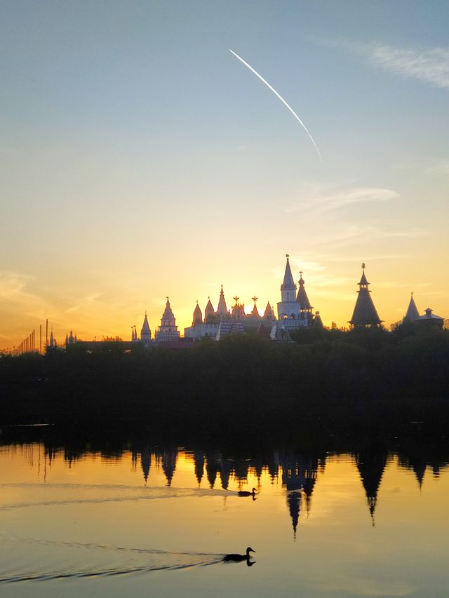 The river is orange from sunset on which ducks swim.  The domes of the castle are visible across the river among the green trees.  The sun is setting behind one of the domes.  The white stripe of the aircraft taking off is visible in the blue sky