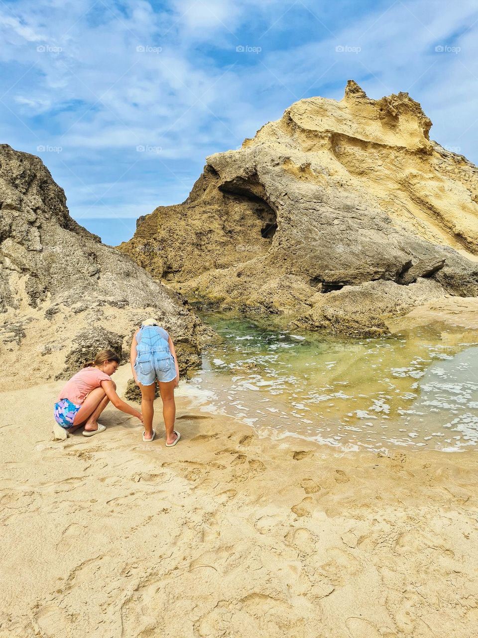 Two young girls playing on the beach next to a small tide pool. it is a beautiful sunny day.