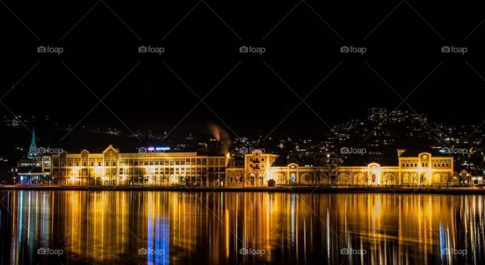 Reflections dance across the water, as the city lights paint the night in vibrant hues