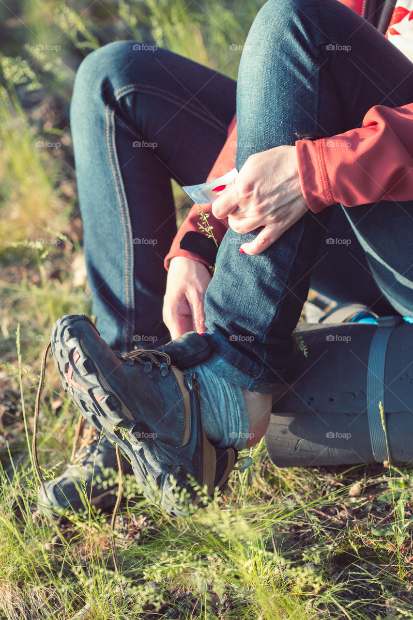 Young woman hiker with backpack taking a shoe off to put a plaster aid on her foot due to tight shoe during summer vacation trip in forest