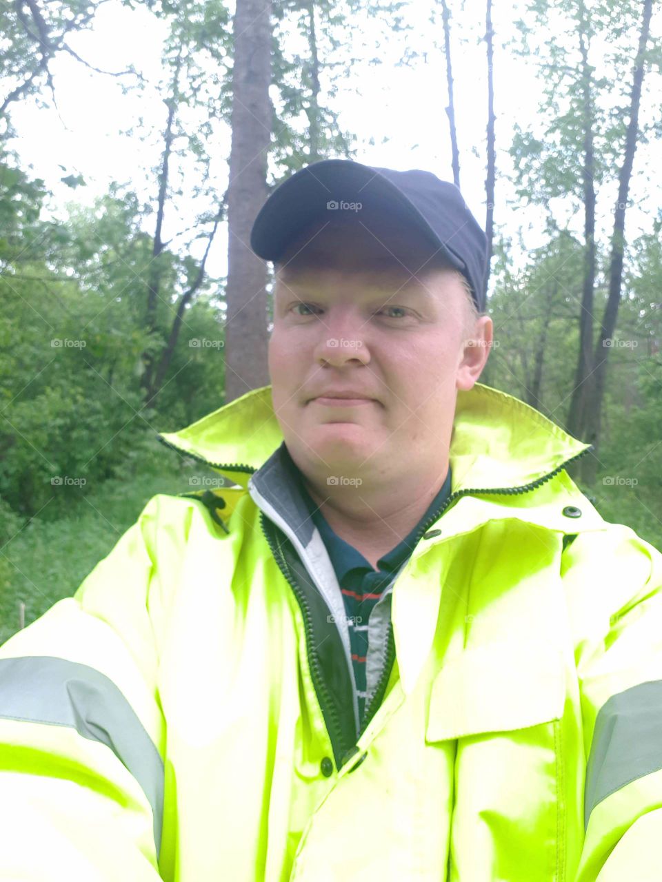 A man in a green jacket and a black cap against a leafy forest
