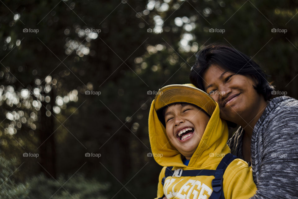 Seasonal outdoor happy family portrait of a young Eurasian kid and his mom hiking in a pine forest wood.Natural setting, the boy is wearing yellow sweater with hood and cap, laughing and the mother smiling, eyes closed