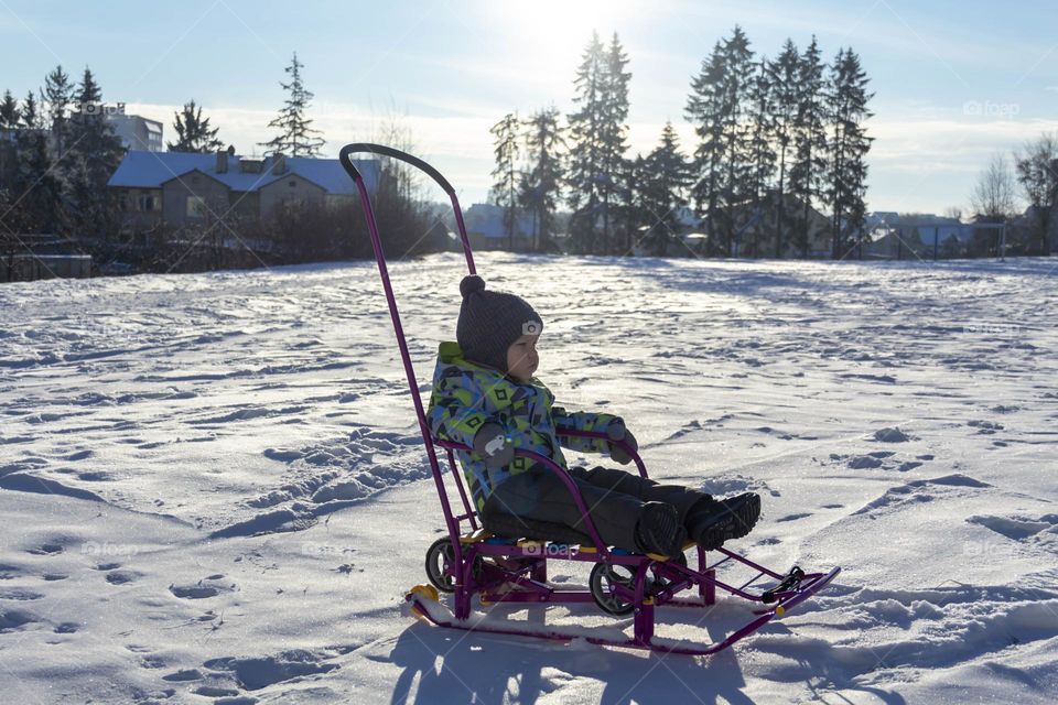 A child with a serious expression on his face in winter clothes jackets, pants, hat and boots in winter on white snow on the street and in the park in nature sledding and playing winter fun.