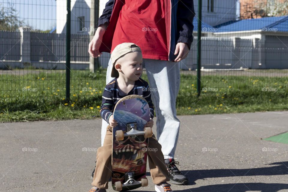 The older brother takes care of the younger, little brother and teaches him his favorite hobby - skateboarding on the sports ground, in the fresh air.